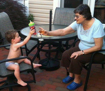 A older woman and a baby are sitting at a patio table. The woman has a glass of wine and is toasting the baby, who is toasting back with a sippy cup.
