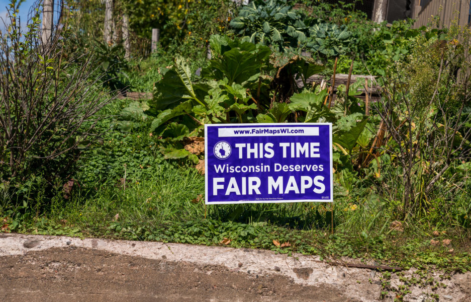 a purple yard sign stuck into some grass reads, "this time Wisconsin deserves fair maps"