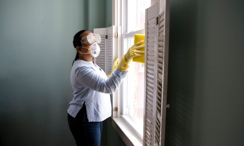 a woman stands at a window, cleaning. she is wearing protective equipment: gloves, goggles, and a mask.