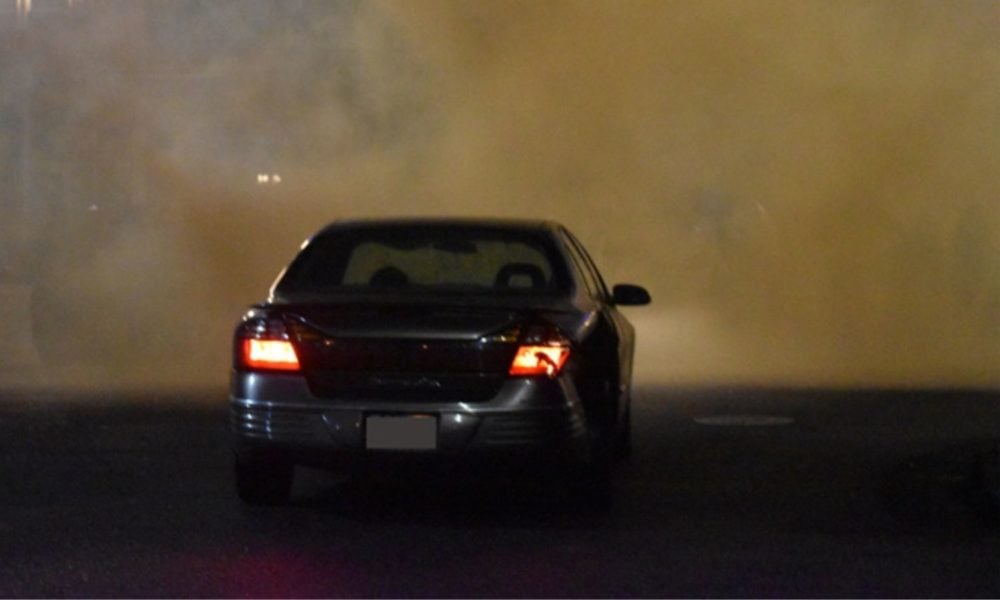 a residential street at night, dark with a few streetlamps in the top left corner. The air is thick with orange and yellow colored smoke / gas that makes it difficult to discern what and who is in the frame of the picture but behind the smoke, although a few silhouettes are peaking through on the right side. The smoke / gas mix is very reactive and dangerous, and so if you were present there, you would be able to feel a chemical burning sensation on any exposed skin. In the center of the photo is a small sedan-style car driving towards the smoke but with brake lights on