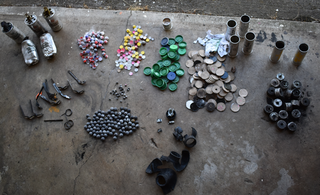 An organized set of piles of chemical weapons items on the ground, which is concrete and asphalt. There are (starting in the top left corner, working clockwise) metal grenades, pepper rounds, FN303 metal/powder rounds, plastic caps and paper wad for shot shells, shot shell casings, chemical inserts for the shot shells, exploded rubber grenades, rubber bullets covered in powder, grenade pins and clips. There are a variety of colors, specifically among the powder rounds (red, yellow, pink), most everything else is a version of grey/black. 