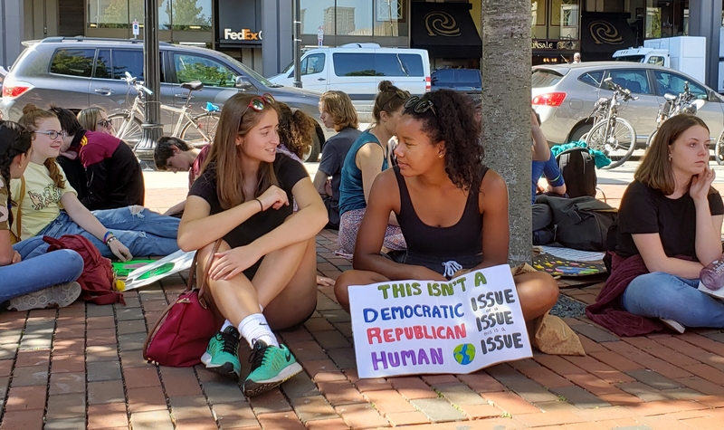 Two youth protesters resting in the shade with sign that implies climate change is not a partisan issue. 