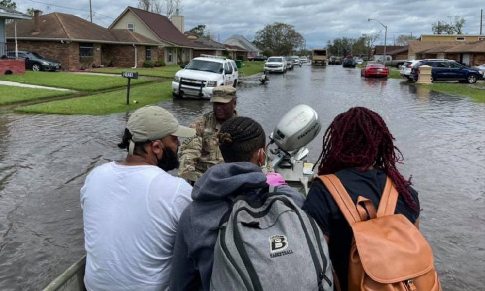 Three people sit in a small motorboat staffed by a member of the National Guard, evacuating them out of a flooded neighborhood