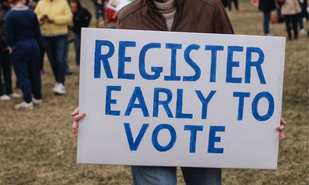 a white man in a brown leather jacket holds up a sign that says 'register early to vote'