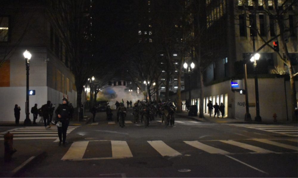 Image of a dark intersection in Portland, OR, with Department of Homeland Security officers dressed in uniforms and wearing masks as they walk away from plumes of smoke behind them.