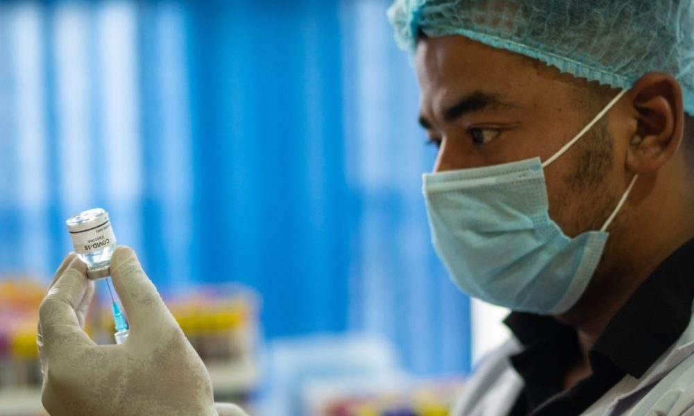 A doctor or nurse wearing a face mask and hair net sticks a hypodermic needle into a vial of COVID-19 vaccine