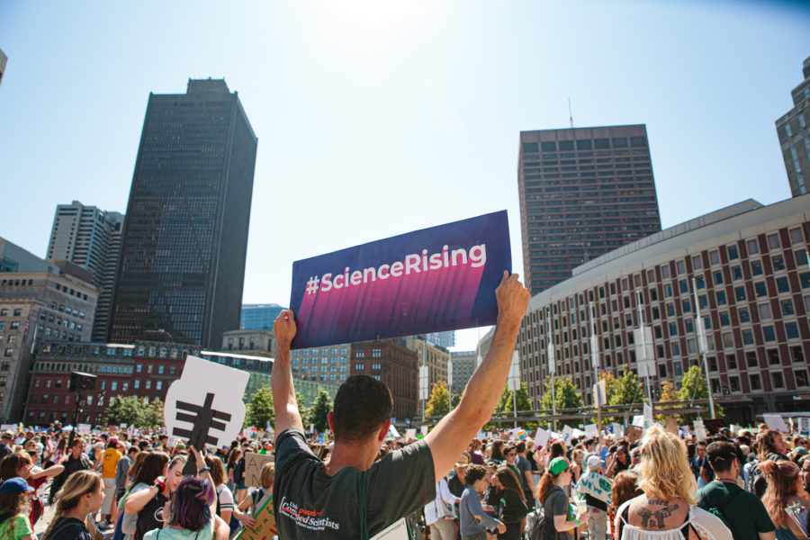 A man holding a purple sign that says #ScienceRising is standing in a crowded square with many other people, some of them also holding signs. There are skyscrapers in the background.