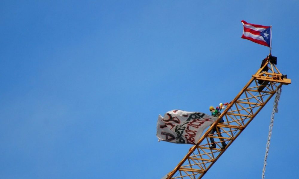 On November 14, 2007, Puerto Rican activist Tito Kayak climbed the construction cranes at the Paseo Caribe project in San Juan, Puerto Rico, and remained perched on a construction crane at the site of a new resort in San Juan for one week, protesting its construction. This is a photo of a blue sky with a yellow construction crane in the foreground, with activist Tito Kayak standing on the crane and a Puerto Rican flag flying at the top.