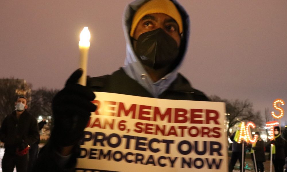 A person wearing a face mask, hat, and hoodie holds a candle and a sign reading "Remember Jan. 6, senators. Protect our democracy now," at a candelight vigil in Washington, DC, after the attack on the US Capitol