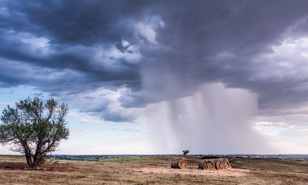 A pastoral scene of some hay bales and a scrubby tree against a flat field, with an ominous looking tornado-like cloud gathering overhead.