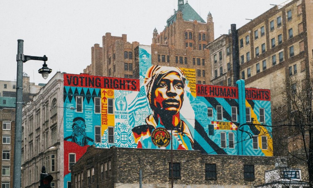 A cityspace of buildings in Milwaukee, WI, with a Shephard Fairey mural on the side of one building reading, "Voting rights are human rights"