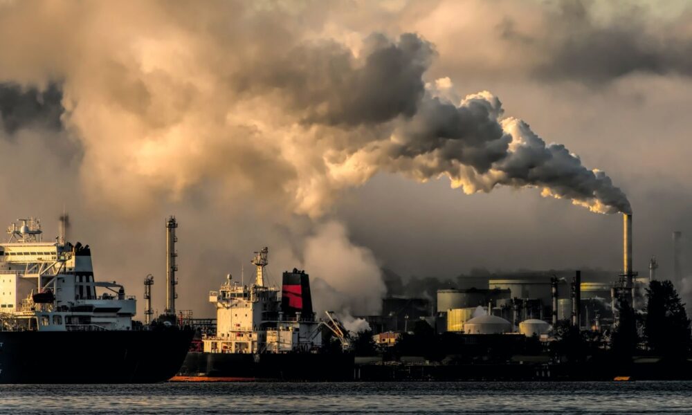 A smokestack belches a cloud of dirty smoke at a port, with ships also leaving clouds of exhaust in the foreground