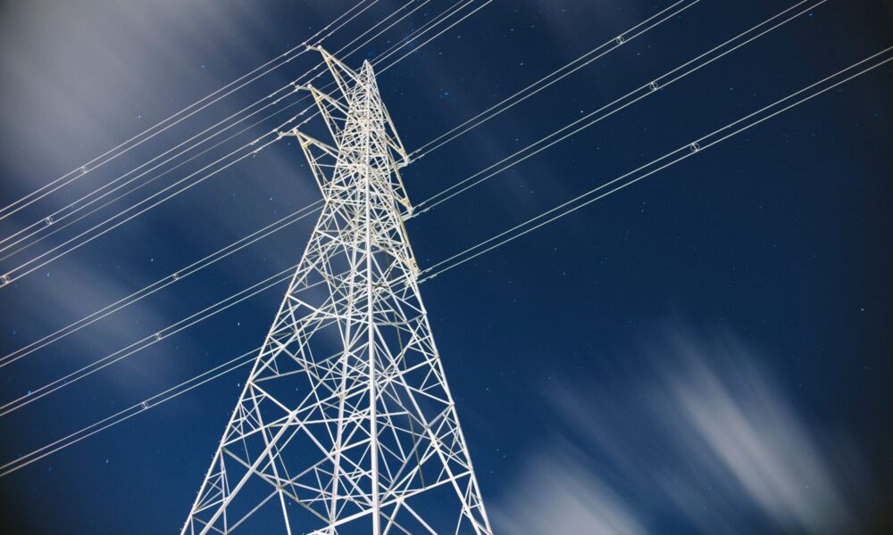in this low-angle photo of a power transmission tower, a blue sky is visible and in the foreground, a white power transmission tower connected to power lines