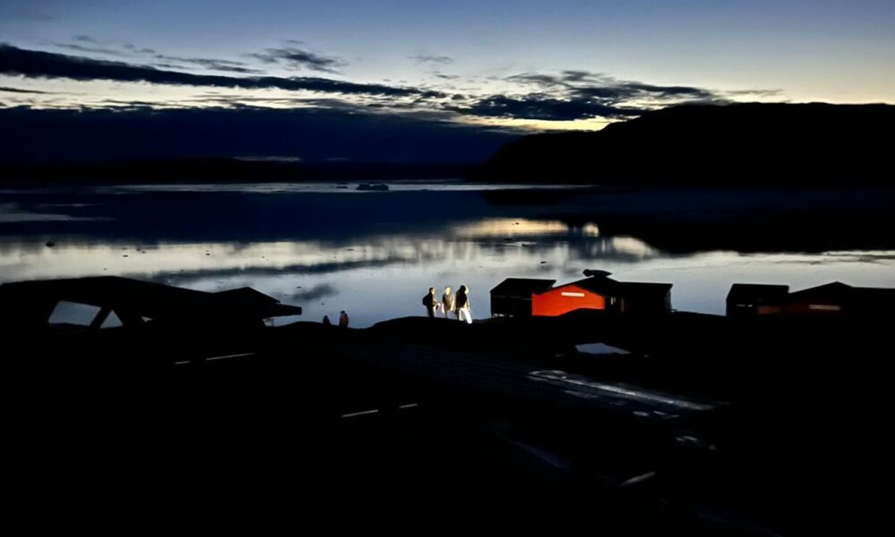 From a distance in twilight, three women stand at the entrance to a cabin with flashlights. A lake is visible in the background.