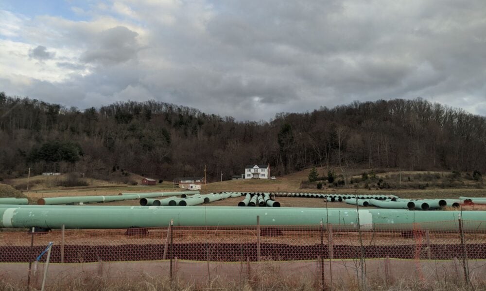In the foreground, a series of green pipes intended to carry natural gas sit atop a hilly landscape in VA or WV, with a house visible in the background