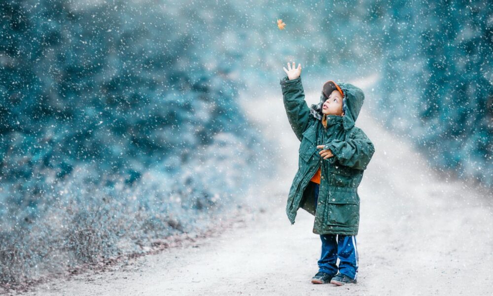 Child reaching out to touch falling leaf on snowy day.