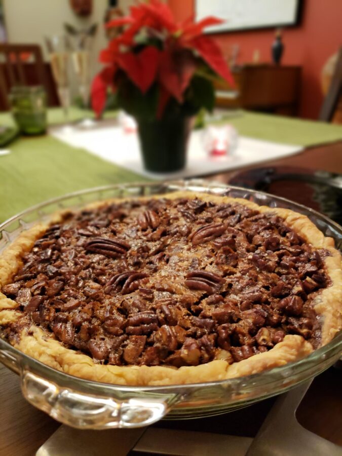 photo of a freshly baked pecan pie in the foreground, on a dining table with a poinsettia plant centerpiece in the background