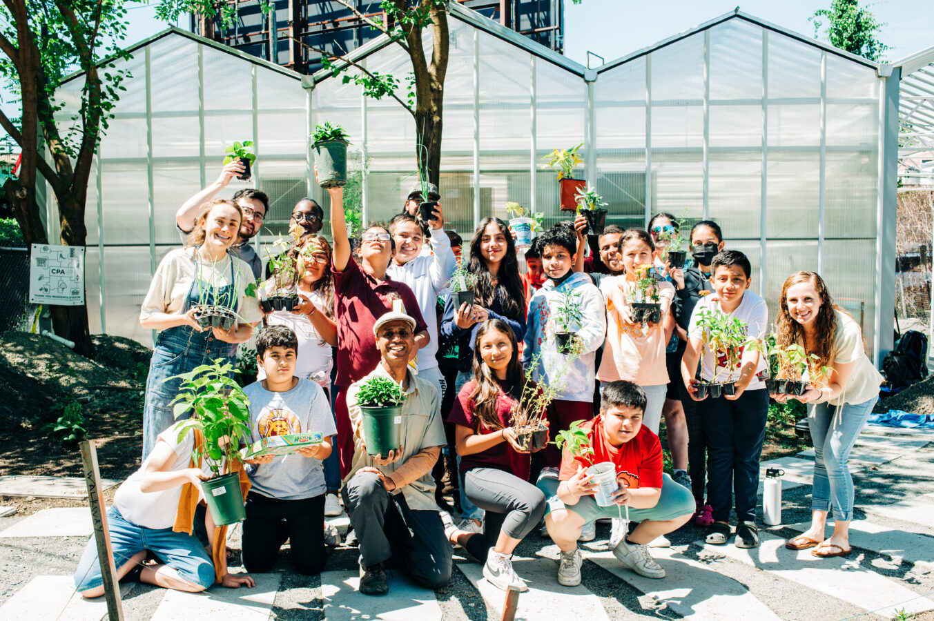 photo of a group of children posing with the director of Eastie Farm in front of the farm's greenhouse in East Boston, MA; everyone is holding up a plant
