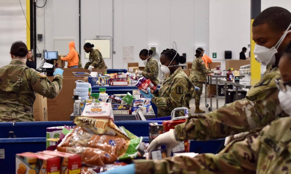 photo of soldiers in camouflage uniforms and masks at a long table filling many crates with food supplies