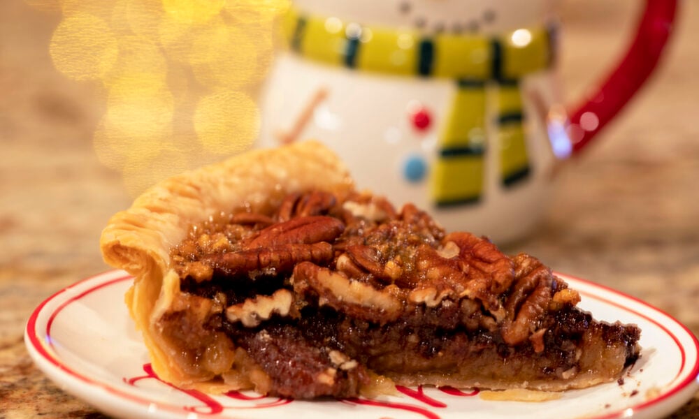 photo of a slice of pecan pie on a red and white plate in the foreground, with a snowman mug in the background