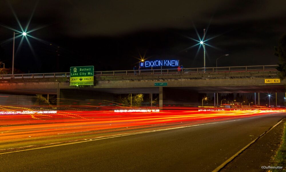 cars' headlights and taillights blur on a Washington-state highway, under an overpass with LED lights on it spelling out #ExxonKnew