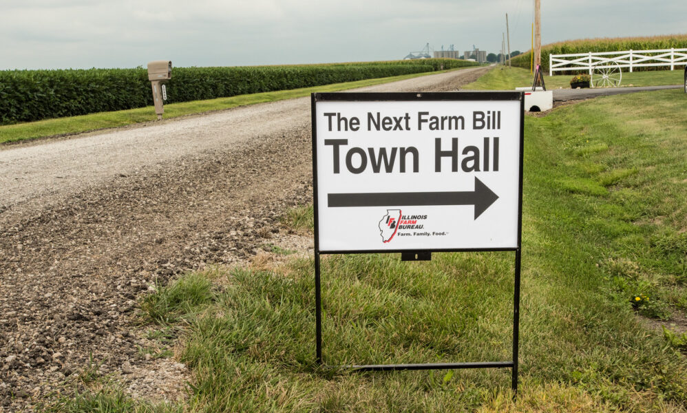 photo of a sign next to a rural road. It reads "The Next Farm Bill" at the top, and "Town Hall" below, with an arrow pointing away from the road.