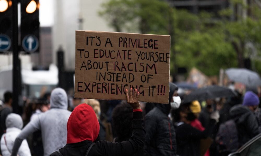 a person's hand holds a sign at a protest that reads "It's a privilege to educate yourself about racism instead of experiencing it!!"
