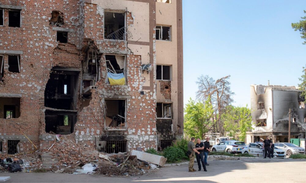 Soldiers converse in front of a brick apartment building showing heavy damage from bombing in Kyiv, Ukraine, with another bombed-out building in the background.