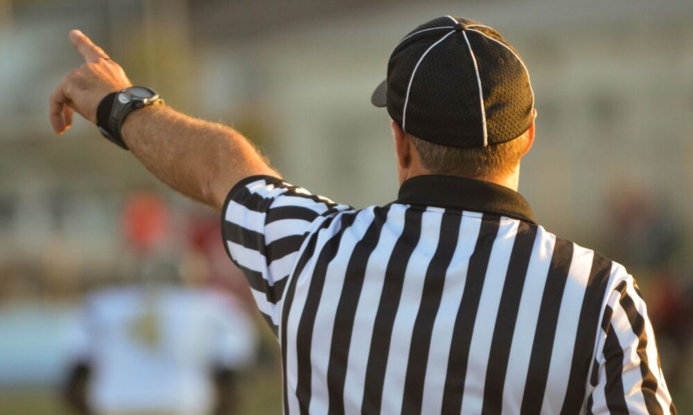 a referee in a black & white striped shirt is pictured from behind, pointing at a call on the field