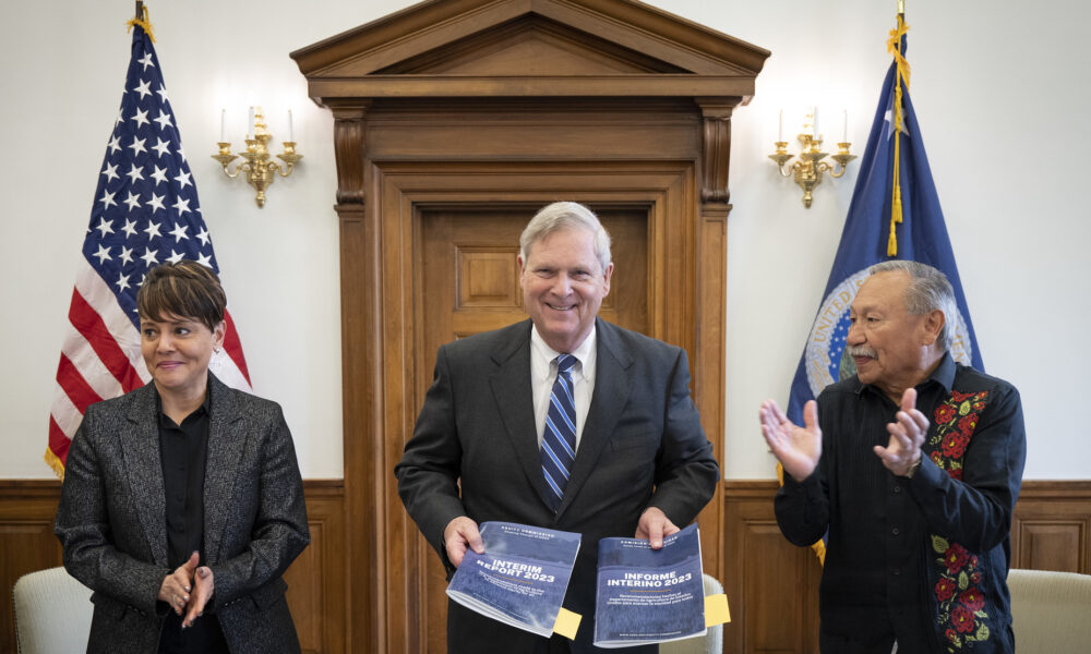 photo of a smiling US Secretary of Agriculture Tom Vilsack holding copies of the 2023 interim report by the USDA Equity Commission; he is flanked by one female and one male member of the commission who are both applauding; the US flag and the USDA flag are in the background