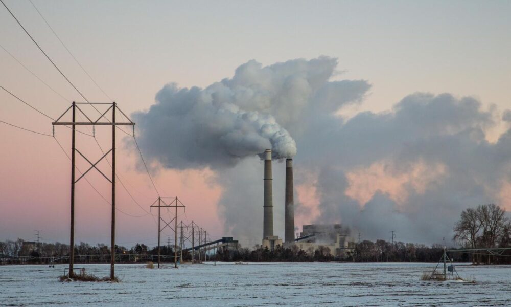Clouds of pollution billow over two smokestacks, part of a power plant in Minnesota, against the backdrop of a pink sky and blue water
