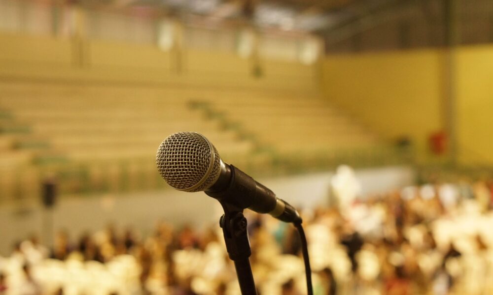 Against a cheery yellow-toned background of people and seats, in an auditorium of some sorts, a black microphone is foregrounded, looking as though it's waiting for you to say something into it