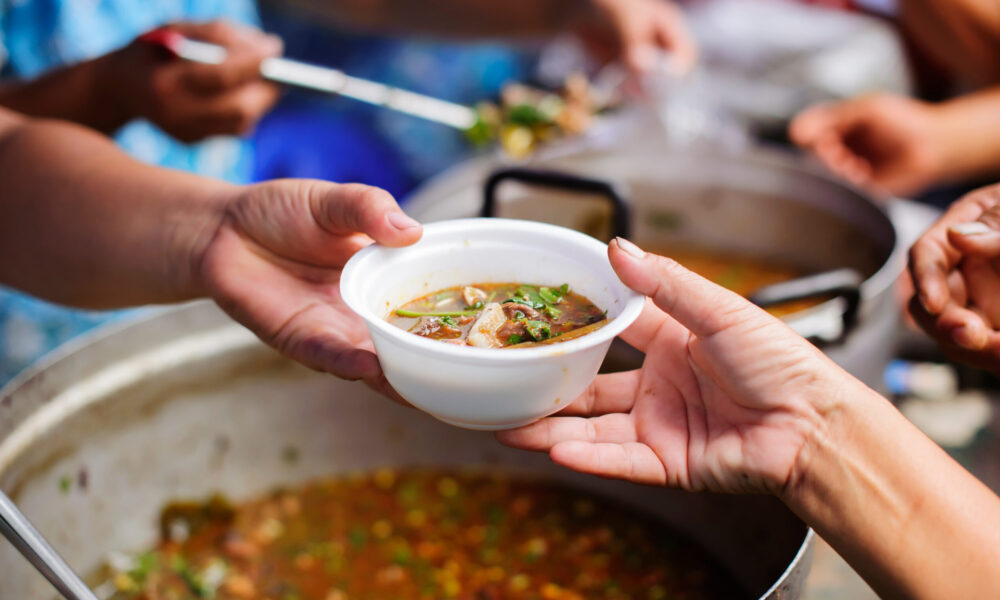 close-up photo of one hand reaching out with a bowl of soup and another hand taking the bowl; in the background are other hands dishing out soup and other hands ready to accept the soup