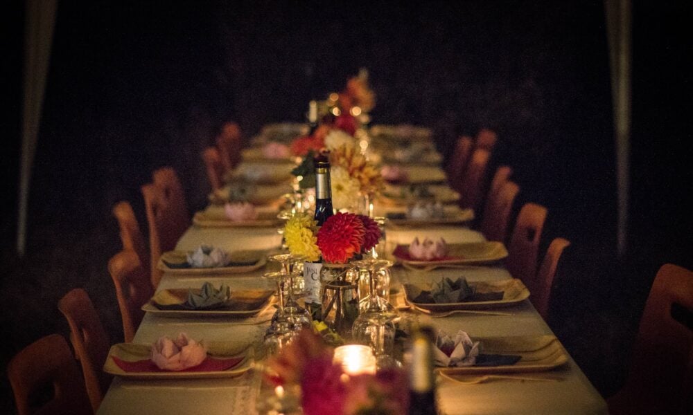 photo of a long dining table in a dark room, ready for dinner with candlelight, flowers, wine, tablecloth and place settings; the chairs are all empty waiting for guests to arrive