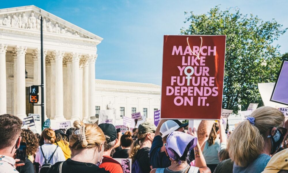 A group of people hold signs at a protest in front of the US Supreme Court building. In the foreground, one sign reads, "March like your future depends on it," with the O in 'your' and T in 'future' aligning to create the symbol for woman.