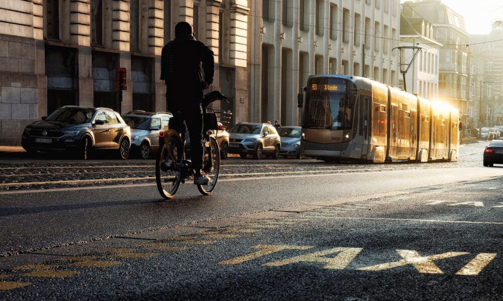 A person rides a bicycle down a city street, past in-ground trolley tracks, a tram, and a taxi stand with cars lined up.