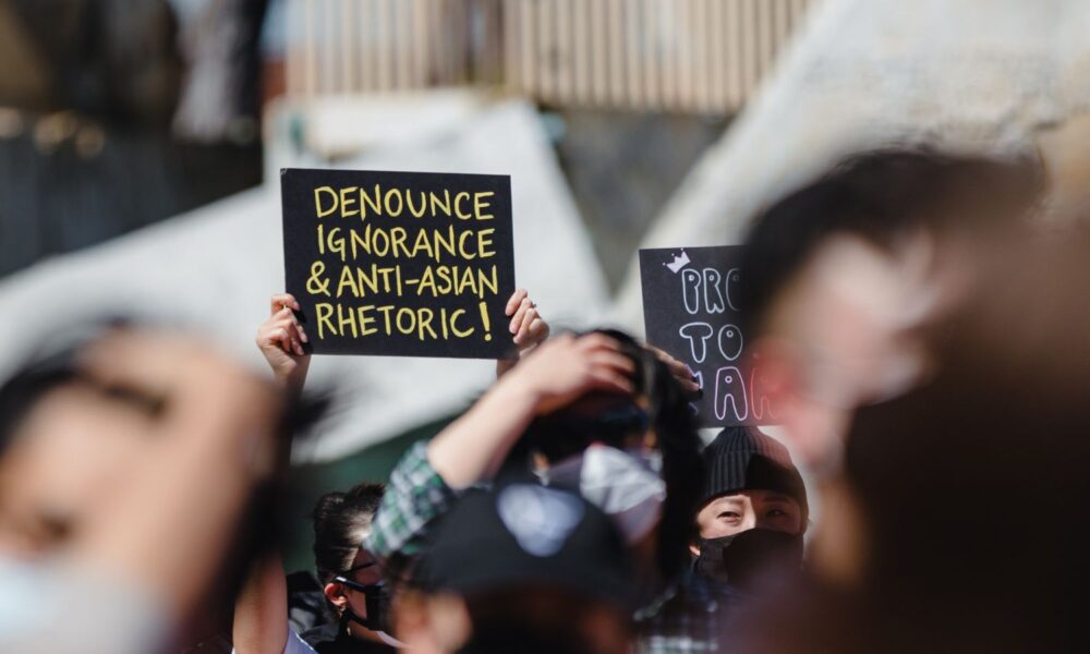 At a 2021 protest in San Francisco, the Rise Up With Asians Rally & March, a person in a crowd holds a sign in focus that reads, "denounce ignorance and anti-asian rhetoric!"