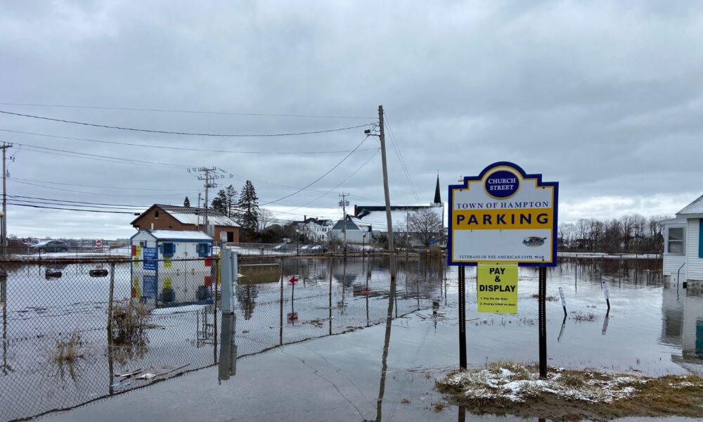 A municipal parking lot in the town of Hampton, NH, is completely flooded, with several buildings nearby whose foundations are obscured by flooding.