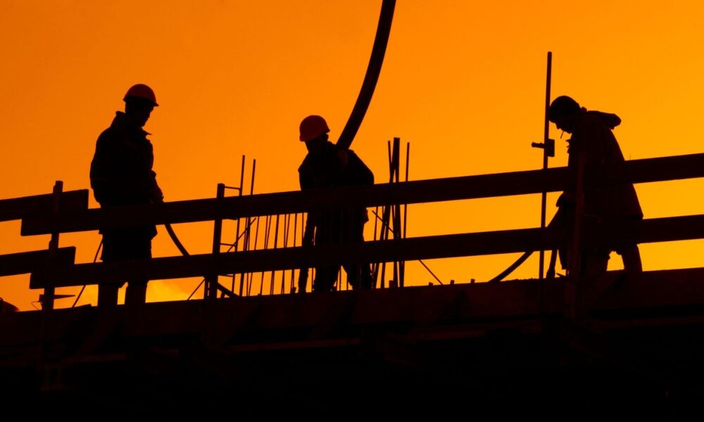 Against a backdrop of an orange sky, a group of construction workers in silhouette are standing near a fence