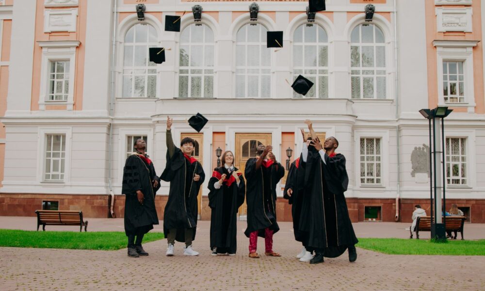 a diverse group of students in graduation gowns stand in front of an academic building, looking up at their graduation caps that they've tossed into the air