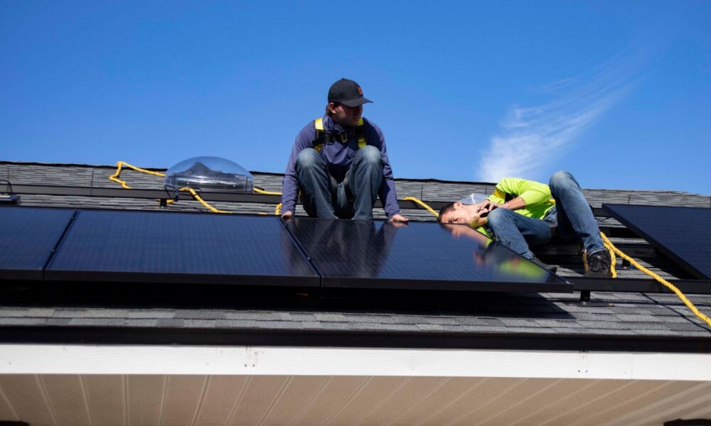 Two white men work to install a solar panel on the roof of a home in St. Augustine, Florida, under a bright blue sky
