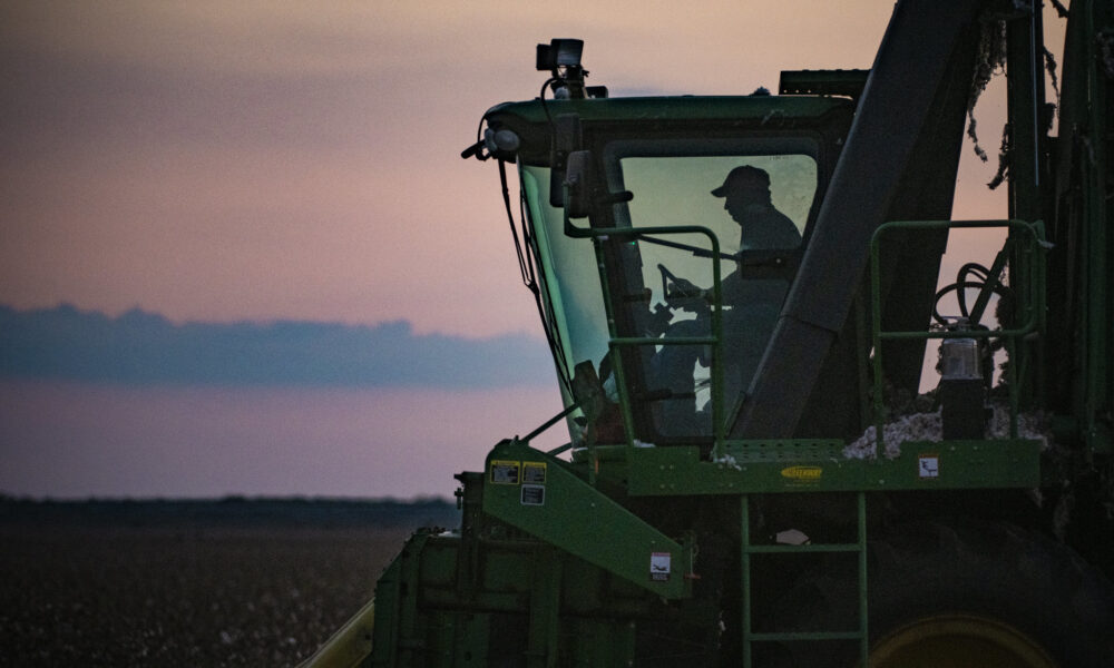 photo of a farmer in the cab of a harvester, driving it through a field at dusk, with the last light of the sun in the background