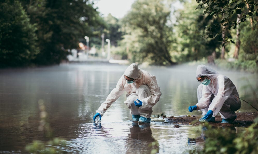 photo of two scientists in protective suits squatting over the surface of a shallow stream in order to take water samples