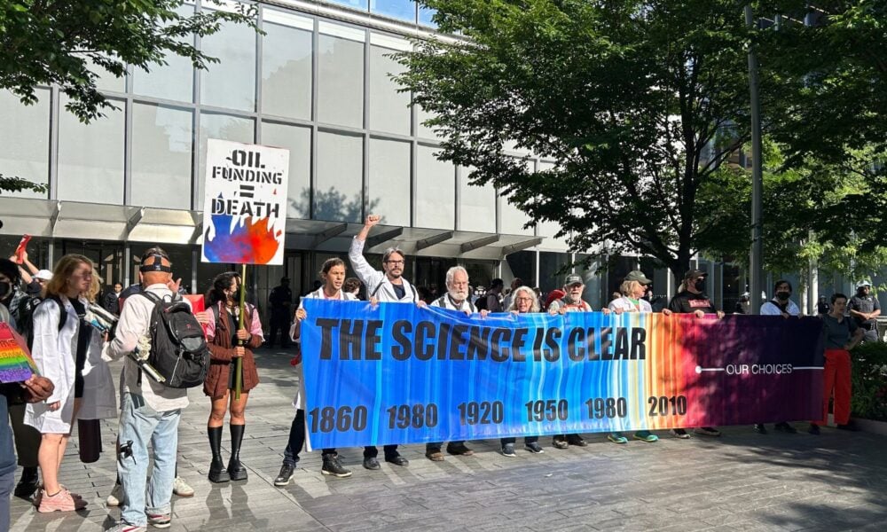 A group of scientists stand in front of Citigroup on Wall St. during the Scientists Speakout Day in June 2024. They hold a banner showing the global average temperatures from 1860 via a color graph that becomes much redder after 2010.