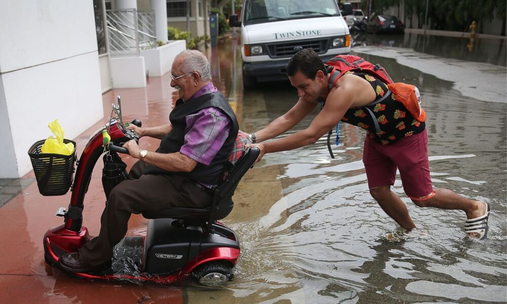 Man pushes man in a motorized scooter across flooded sidewalk.