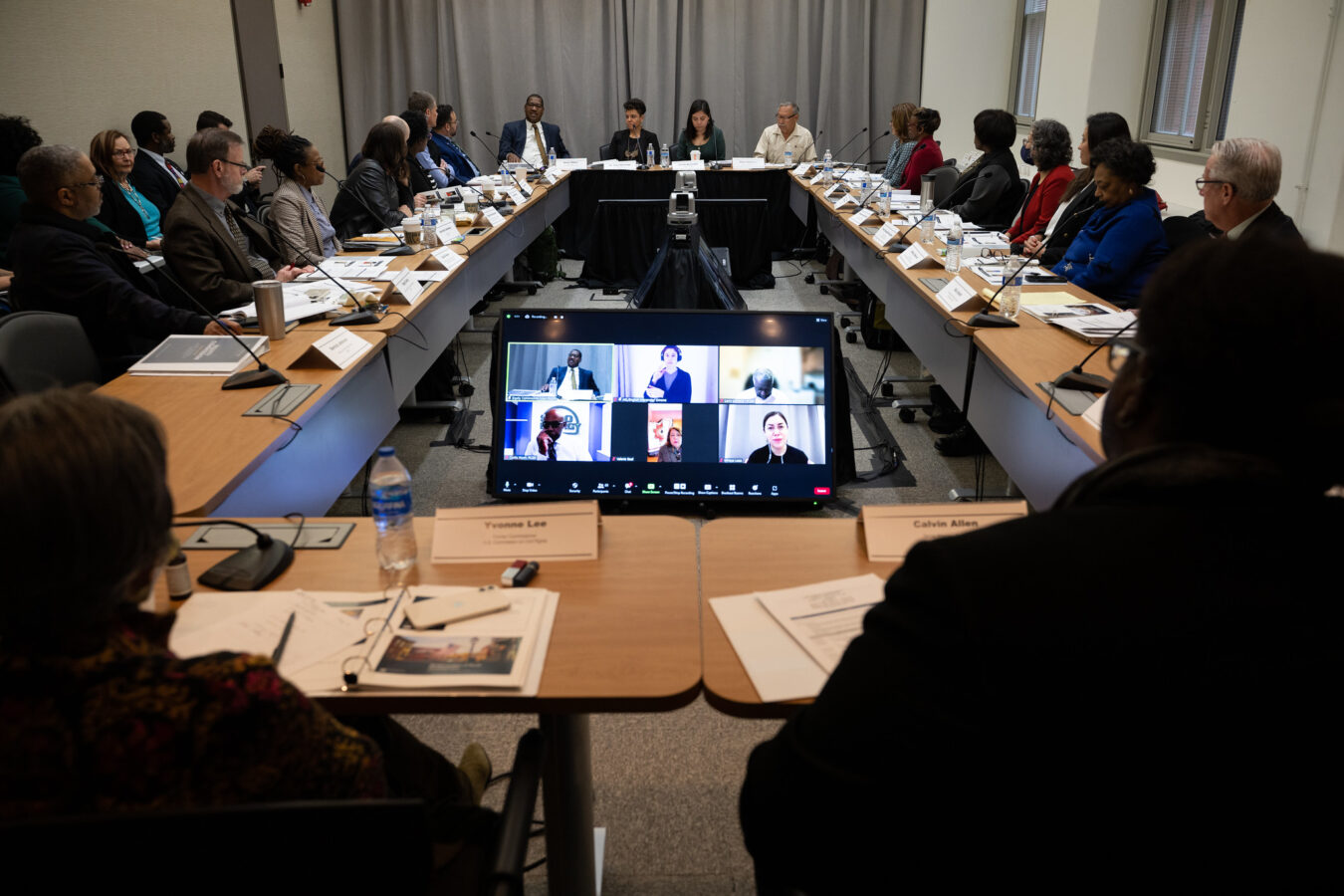 photo of the January 31, 2023 meeting of the USDA Equity Commission in Washington, DC; a video screen showing remote participants is in the foreground and there are rows of commission members seated on either side facing each other, with notebooks and microphones for each person