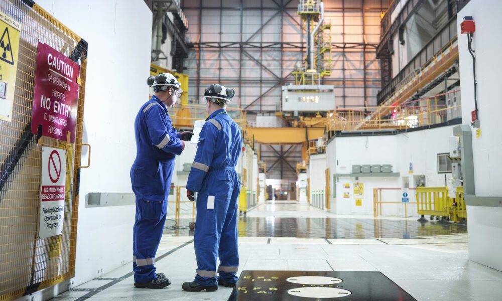 photo of two men in blue jumpsuits and hard hats talking inside the cavernous reactor hall of a nuclear power plant; there are caution signs and a radioactive symbol on the wall next to them