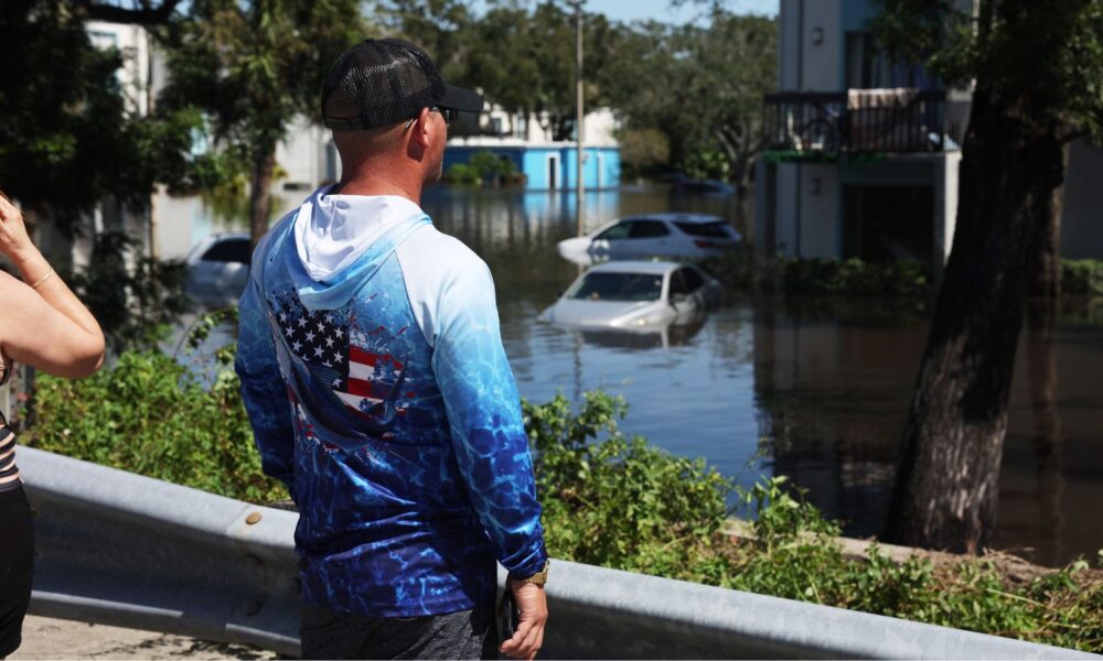 A person wearing an American flag hoodie surveys a flooded apartment complex and submerged parking lot in Clearwater, FL, after Hurricane Milton in 2024.