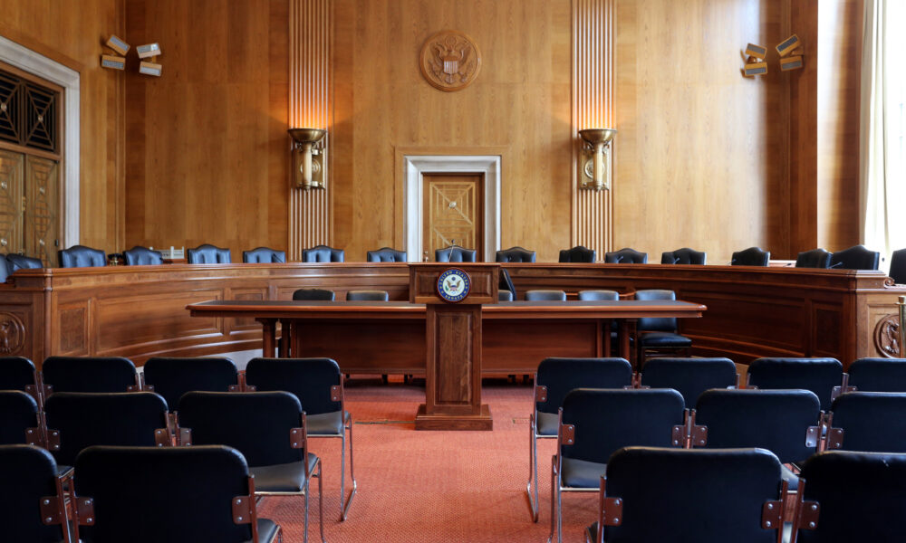 photo of an empty US Senate hearing room, with a row of padded blue chairs on a raised dais facing out toward multiple rows of metal chairs for visitors