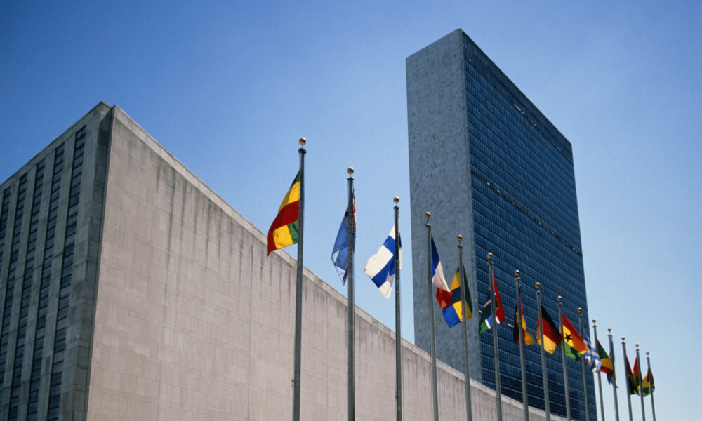 photo of the United Nations headquarters in New York City; in the background, the tower rises against a blue sky and, in the foreground, a row of various nations' flags flutter in the breeze